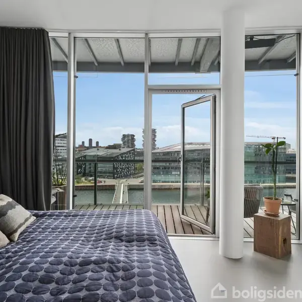Soveværelse med dobbeltseng i forgrunden, åbne glasdøre fører til en balkon med udsigt over en kanal og moderne bygninger. Rummet er lyst med sorte gardiner.