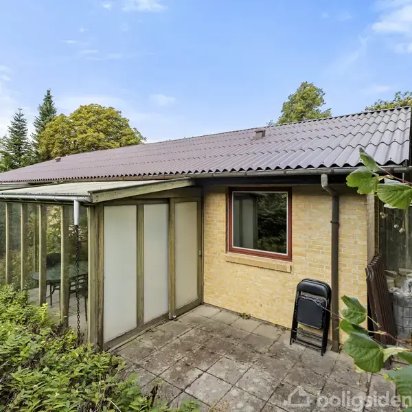 Huset med gul mursten og mørkt tag står i en frodig have. En overdækket veranda på venstre side er omgivet af grønne planter og en stenbelagt terrasse med en stol.