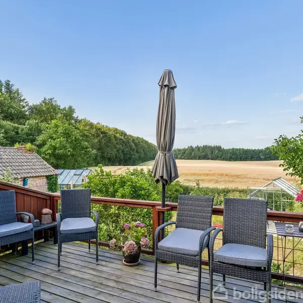 Terrasse med sorte kurvemøbler og sammenklappede parasoller står roligt. Omkranset af grønne træer og en gylden mark i baggrunden, giver en fredfyldt udsigt over det naturskønne område.