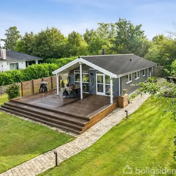 Et gråmalet hus med terrasse står i en frodig have. En sti fører op til terrassen, hvor der er havemøbler. Området er omgivet af træer og grønne buske.