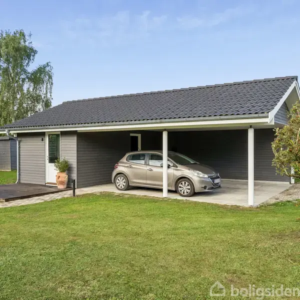 En bil holder parkeret under en carport ved et mørkt træhus. Omkring huset er der en græsplæne og nogle buske. Himlen er klar med få skyer.