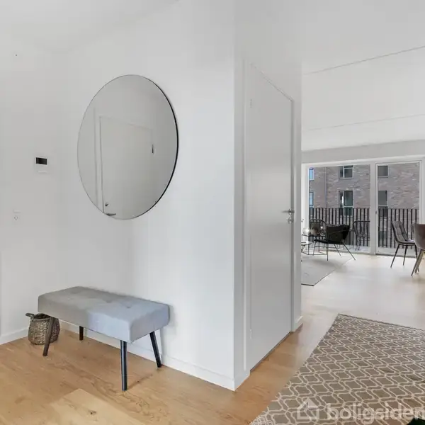 En lys entré med en rund vægspejl og en grå bænk. Til højre ses et åbent område med spisebord og stole ved en altan.