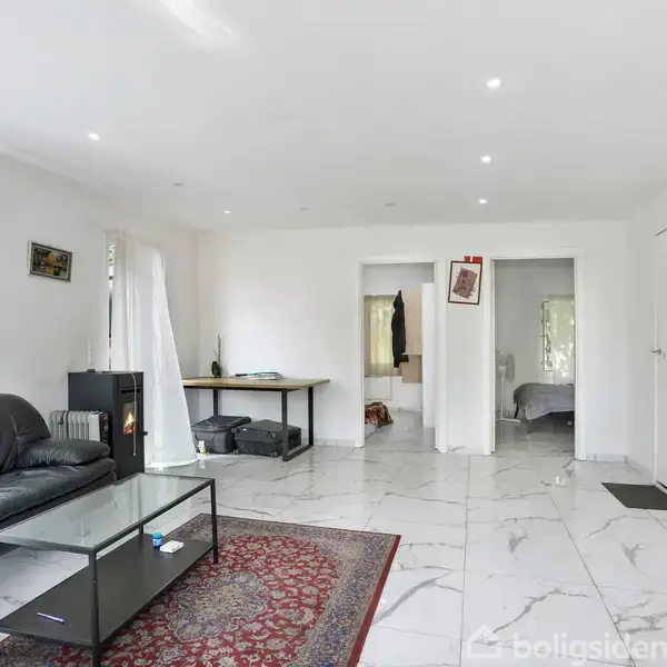 Stue med sort lædersofa og glasbord på rødt tæppe. Handling mangler, da rummet er stille. Lyse vægge og gulvfliser skaber moderne, minimalistisk atmosfære. Indgang til soveværelse ses i baggrunden.