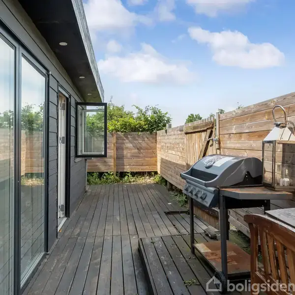 En træterrasse med en bordgrill står op ad en træbeklædt væg. Der er et åbent vindue og et bord med stole i et aflukket udendørsområde.
