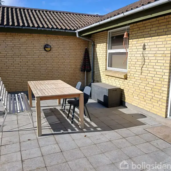 Et træbord står på en flisebelagt terrasse sammen med stole og en bænk. Omkring er gule murstensvægge, et vindue og en dør, alt under et tegltag.