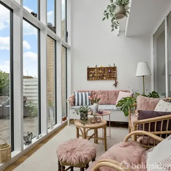 En lys stue med store vinduer viser en hyggelig siddeplads med rottingsmøbler og lyserøde puder. Planter dekorerer rummet. Udenfor er der en terrasse med cafébord og stole.