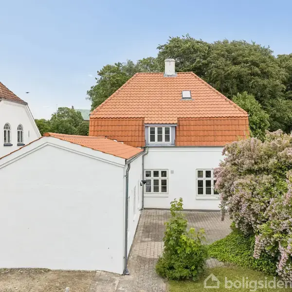 Hvidt hus med rødt tegltag, stående i rolig haveomgivelse. Huset har flere vinduer og en mindre terrasse. Omringet af andre huse og grønne buske.