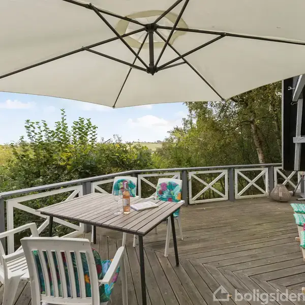 En terrasse med et bord og stole langs et hvidt rækværk. En stor parasol skygger området. Træer og grønt landskab omringer terrassen. Hynder ligger på stolesæderne.