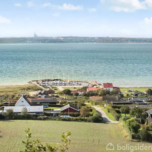 Hus ved kysten omgivet af grønne marker og træer. Foran er en marina ved havet med både, og i baggrunden ses en anden kystlinje på den modsatte side af vandet.