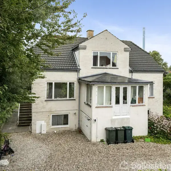 En lys murstensvilla med sort tegltag står stille i en have omkranset af grønne træer. Foran huset er en grusbelagt indkørsel med tre affaldscontainere.