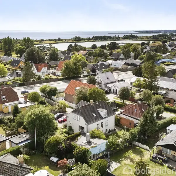 Huse ligger i en grøn forstad, omgivet af træer og haver, tæt på en kystlinje. Røde og gule tagsten kontrasterer mod den blå himmel.
