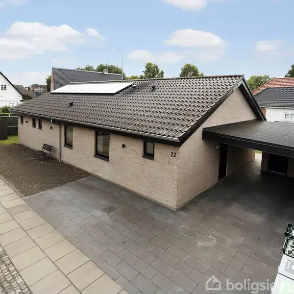 Et enkelt etplanshus med fladt tag og lyse mursten står på en velholdt flisebelagt indkørsel. Der er en carport til højre og grønne buske i baggrunden. Nummer 22 vises på husets facade.
