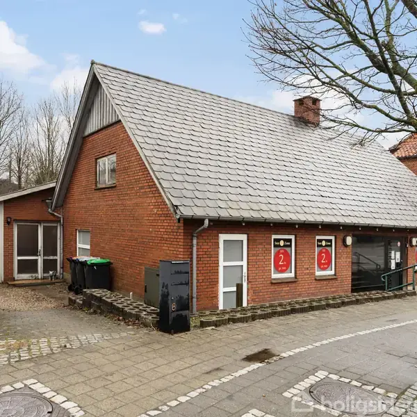Rødt murstenshus med gråt tag og rampe ved indgangen. Fire plakater med teksten "2" i vinduerne. Til venstre ses en garage. Bar træer og fortov i forgrunden.