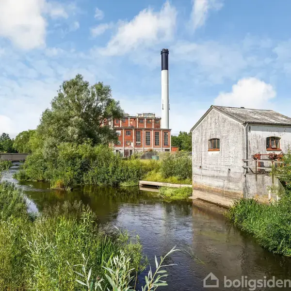 Vaskemaskine og tørretumbler er stablet i et hvidt badeværelse med sort flisegulv. Ved siden af er et toilet og en håndvask med et spejl og en vase med blomster.