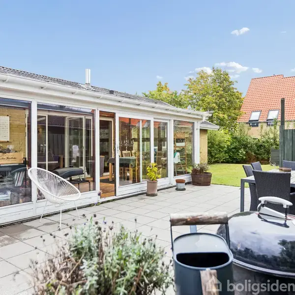 En terrasse med havemøbler og grill står foran et hus med store glaspartier. I baggrunden ses en græsplæne og haveomgivelser med træer og buske.