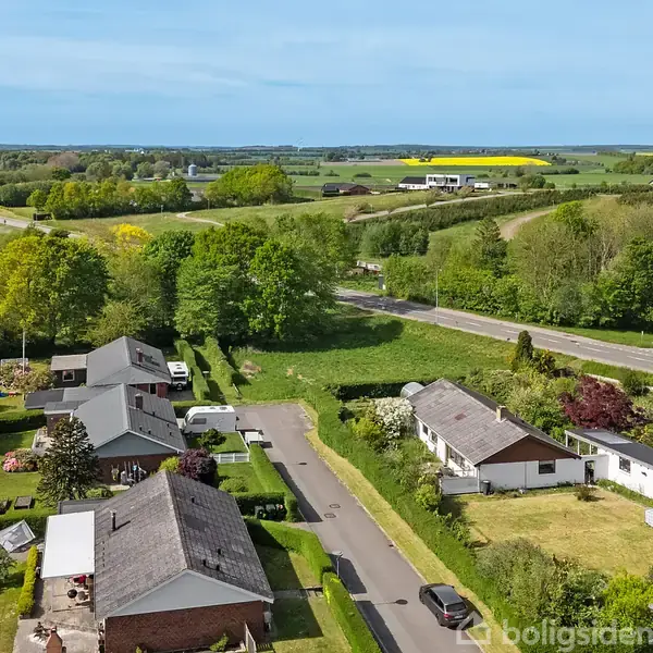 Huse står stille langs en vej, omgivet af grønne haver og træer. I baggrunden ses åbne marker og en hovedvej, der snor sig gennem landskabet på en solrig dag.