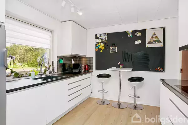 Et køkken med hvide skabe og sorte bordplader. To barstole står ved et rundt bord foran en stor opslagstavle. Et vindue viser en have udenfor.