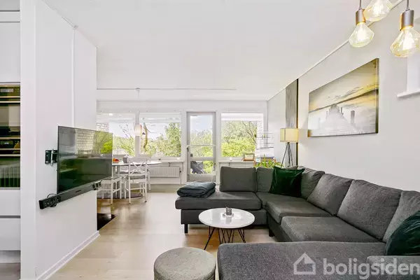 Stor grå sofa i en lys stue placeret ved et moderne sofabord. Et fladskærms-tv hænger på væggen, mens store vinduer i baggrunden giver udsigt til grønt område. En spisestue ses i hjørnet med hvide stole.