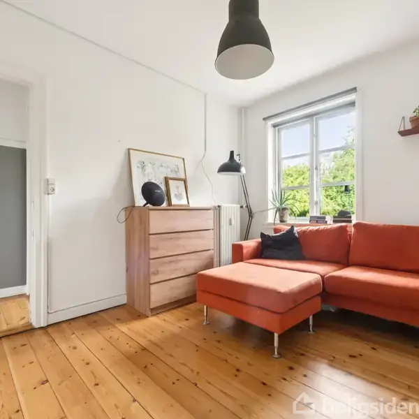 Orange sofa står i et lyst rum med trægulv ved siden af et vindue. En kommode med billederammer og lampe er tæt på, med en hylde over.
