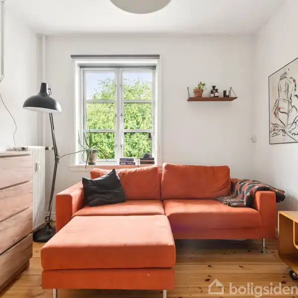 En orange sofa med chaiselong står i en lys stue. Ved siden af er en kommode med en lampe. Et vindue viser udsigt til grønne buske. En TV-enhed står til højre. Ingen tekst synlig.