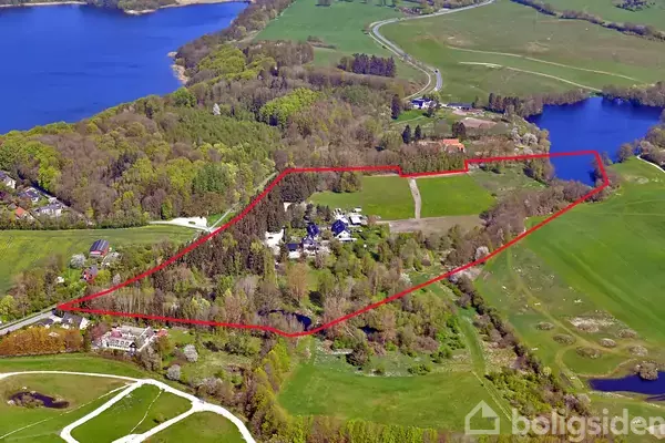 En stor, grøn landgrund omgivet af skov og en sø, afgrænset af en rød linje. I midten ses flere bygninger og veje, samt åbne græsarealer.