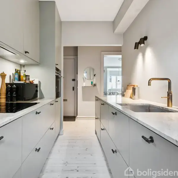 Køkken med grå skabe og hvide bordplader. Køkkenredskaber og flasker står ved kogepladerne til venstre. På væggen til højre er en vask med messingarmatur og sæbe.