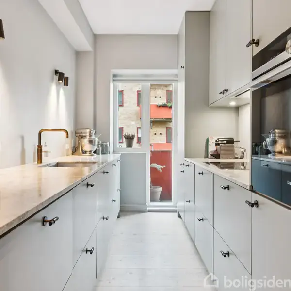 Køkken i moderne stil med grå skabe og hvide bordplader. To vaskebænke står mod hinanden. Vindue for enden giver udsigt til bygninger og balkon.