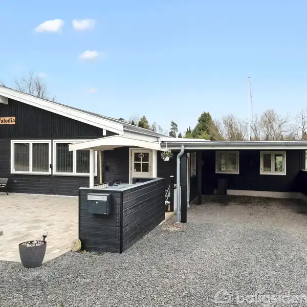 Sort træhus med tilhørende garage står på en grusbelagt grund. Skiltet "Valhalla" er hængt på facaden. Huset er omgivet af træer og græsplæne under en blå himmel.