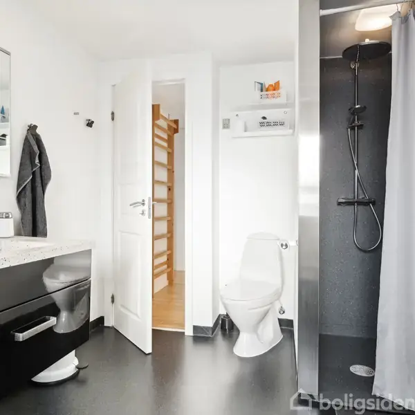 Badeværelse med sort skabsmodul under spejl, toilet ved siden af, bruseniche med grå gardin til højre. Dør åben til andet rum, sort gulv og hvidmalede vægge.