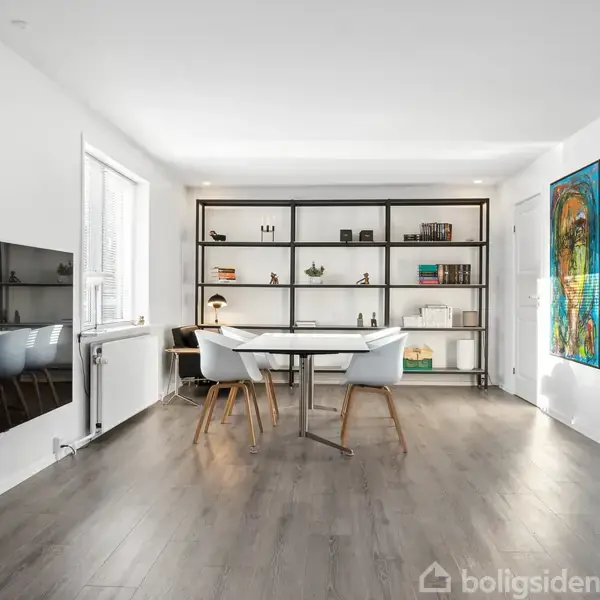 Reol fyldt med bøger og dekorationer står op ad en hvid væg. Rundt bord omgivet af stole, fladskærms-tv monteret på venstre væg, maleri hænger til højre, trælaminatgulv.