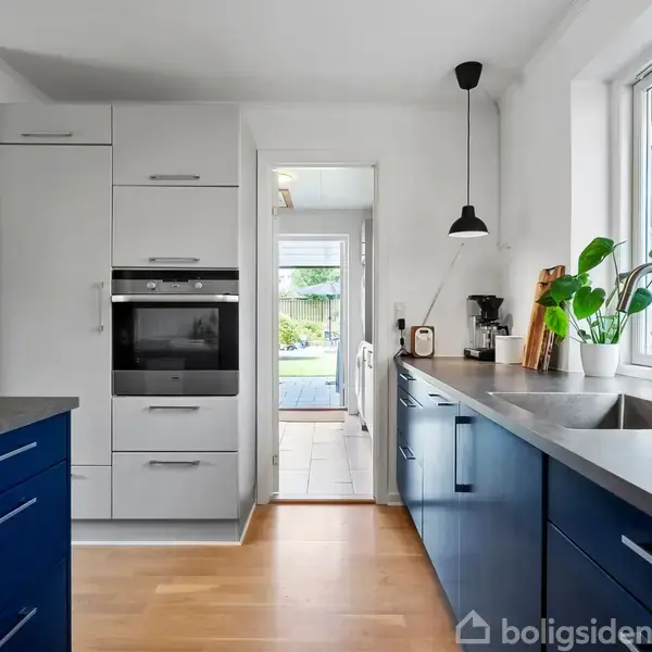 Køkken med blå skabe og lys træbordplade. Ovnen er indbygget i et højskab til venstre. Et stort vindue med planter står ved siden af en vask. En lampe hænger fra loftet.