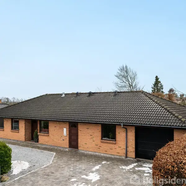 En enkeltstående gul murstensvilla med mørkt tegltag står på en brolagt indkørsel. Omkring huset er der klippet hæk og spredte træer, under en klar blå himmel.
