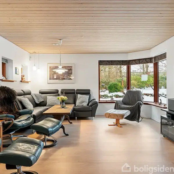 En stue med sorte læderstole og sofa, står roligt omkring et sofabord. Et stort vindue åbner rummet mod en snedækket have. Væggene har billeder og lamper.