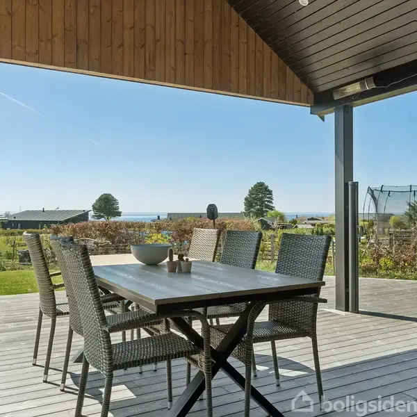 Et udendørs træbord med seks stole står på en overdækket terrasse. I baggrunden ses en have, trampolin og havudsigt under en klar blå himmel.