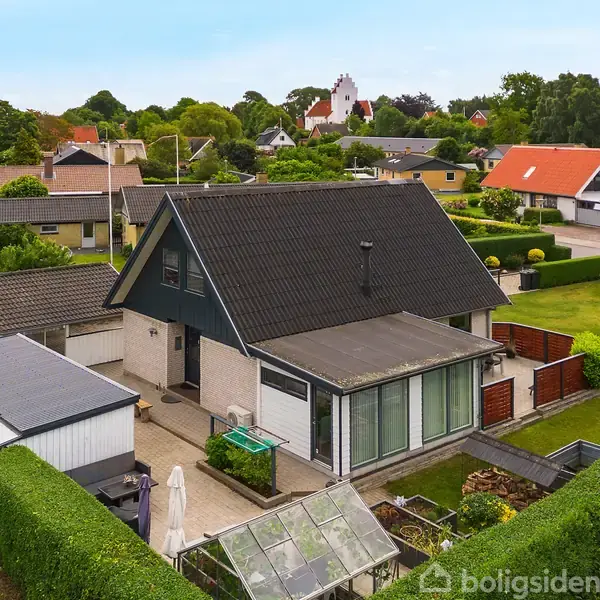 Hus med mørkt saddeltag ligger i forstadsområde omgivet af grønne haver. Der er en terrasse foran huset og flere nabohuse i baggrunden.