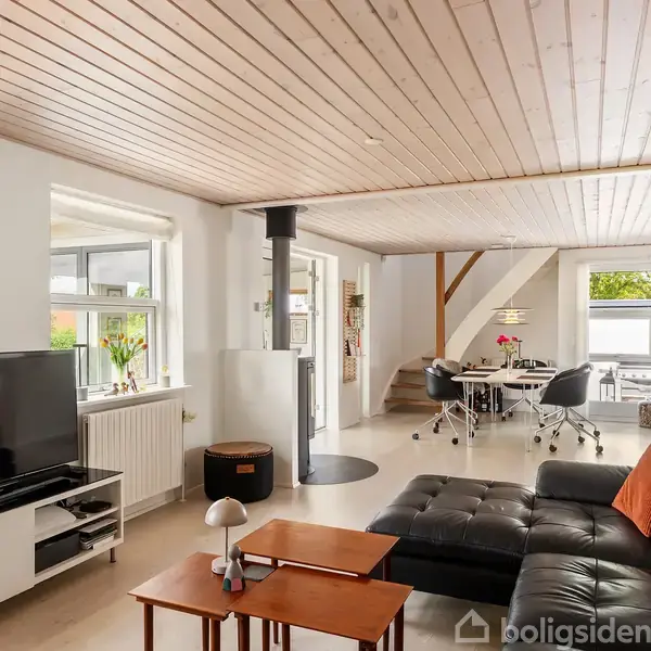 Sort lædersofa står i stue med fladskærms-TV på hvidt skab; spisebord med stole i baggrunden, træloft ovenfor. Naturligt lys strømmer ind gennem de store vinduer.