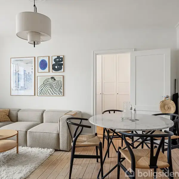 Stue med en beige sofa og lavt kaffebord placeret på et hvidt tæppe. Rundt spisebord med sorte stole. Væggene prydet med forskellige kunstplakater og malerier.