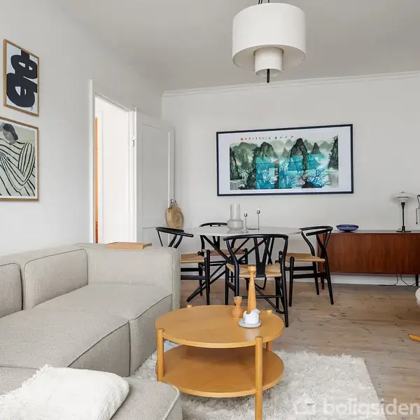 Stue med lysegrå sofa, rund trækistebord og moderne sort spisebord. Væggene har kunstplakater, og et træskab står bag spisebordet. Hængelampe og gyngestol kompletterer rummet.