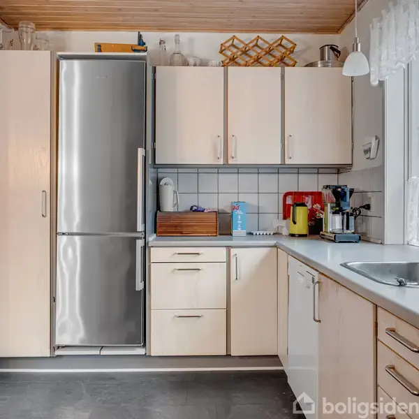 Et køkken med lyse skabe og en stålkøleskab. Bordpladen har kaffemaskine, røremaskine, og brødkasse. Store vinduer til højre lukker lys ind. Væggene er lyse og gulvet er mørkt.