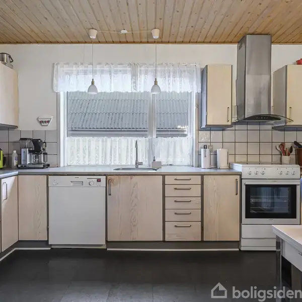 Køkken med lyse træskabe, opvaskemaskine og ovn med emhætte. To vinduer over vasken lader lys ind. Kaffemaskine og køkkenredskaber på bordpladen. Bord med dekorativ krukke i forgrunden.