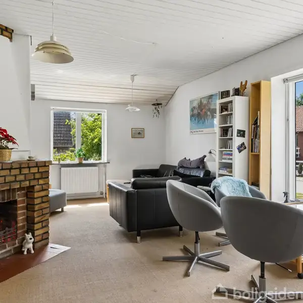 Pejs med levende ild i en lys stue. Møbler inkluderer to grå lænestole og en sort sofa. Vinduer giver udsigt til en solrig have og rødt hus.