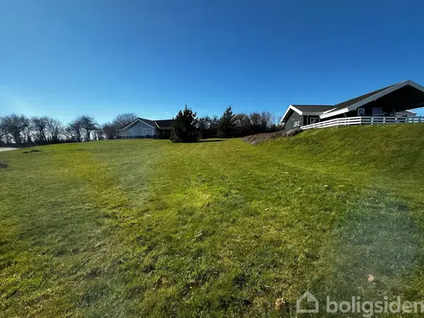 To huse står på en grøn, solbelyst græsplæne med træer i baggrunden; den klare blå himmel forstærker den fredelige atmosfære.