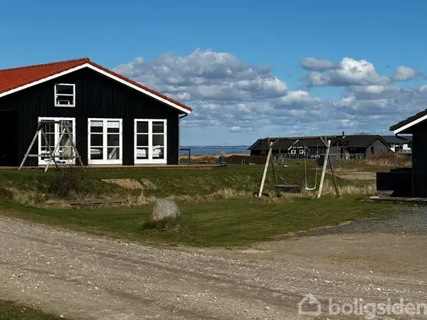 Et sort hus står stille ved en grusvej med gynger uden for. I baggrunden ses flere huse og en blå himmel med skyer.
