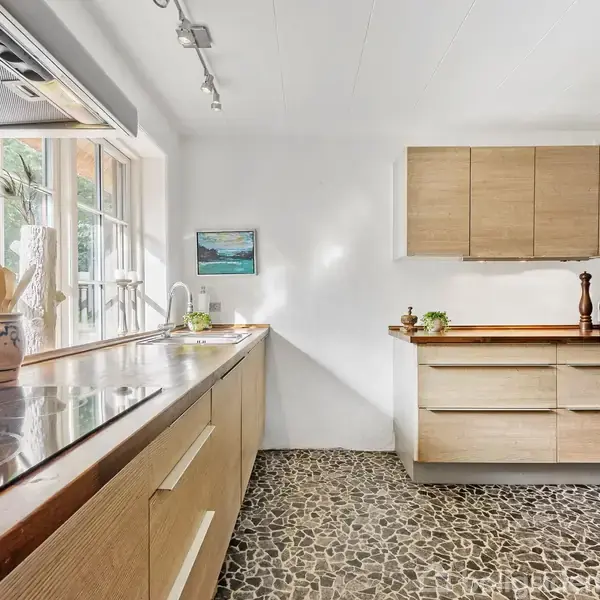 Et køkken med en gråmeleret stengulv, træbordplader og lyse træskabe. På bordet står en krukke med køkkenredskaber. En væg har kunstmaleri, og vinduer slipper naturligt lys ind.