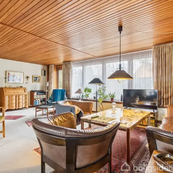 Stuen har sofaer og stole omkring et sofabord, vinduer med tunge gardiner, og hængelamper. Der er tæpper på gulvet og billedkunst på væggene.