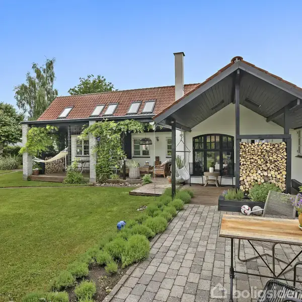 Et charmerende hus med rødt tegltag og glasvinduer har en overdækket terrasse med brændestabel og planter. Omgivet af en velplejet have med græsplæne og buske.