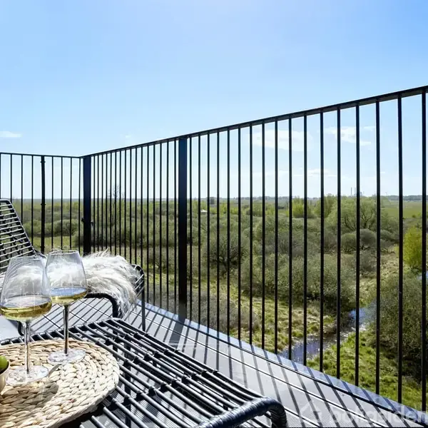 To glas hvidvin og en kurv med grønne druer står på et bord på en balkon. I baggrunden ses en udsigt over et frodigt landskab under en klar blå himmel.