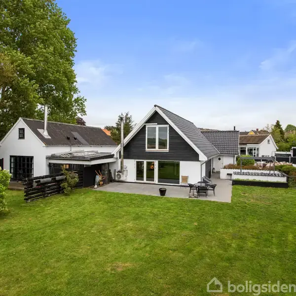 Hus med sort og hvid facade står i centrum i en stor have med grøn græsplæne. Træer omkranser området, og der er en flisebelagt terrasse ved huset.