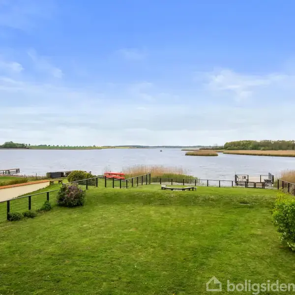 Græsplæne strækker sig mod en bred flod med siv langs kysten; himlen er blå med få skyer. I højre side ses en åben dør, der fører ud.