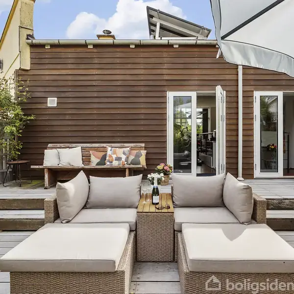 Terrassemøbler står på et trædæk med en hvid parasol over sig. I baggrunden ses et hus med træfacade og dobbelt glasdør, omgivet af grønne planter og puder på en bænk.